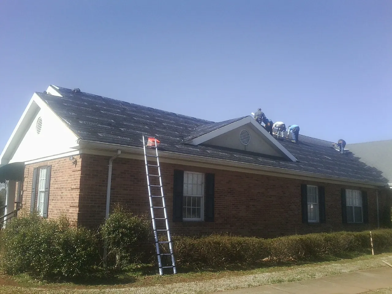 Crew tearing shingles off a brick ranch roof with a ladder set — Emergency Roof Repair work by our crew in Gray
